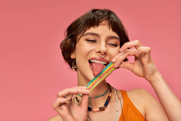 cheerful young woman in her 20s licking sweet and sour candy strip on her tongue, pink background - Photo, Image