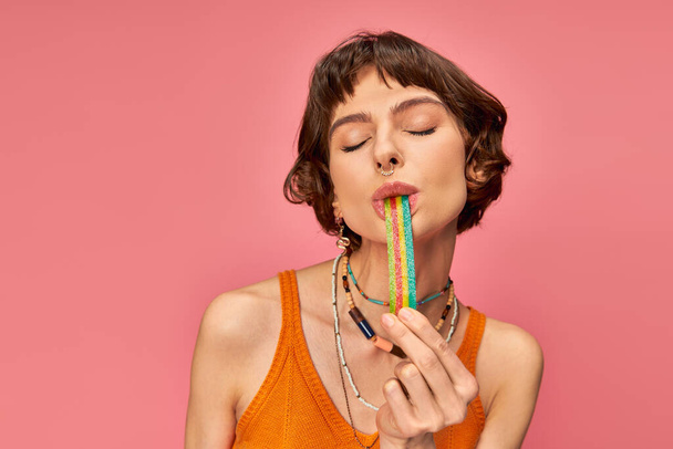 portrait of cheerful young woman in her 20s biting sweet and sour candy strip on pink background - Photo, Image