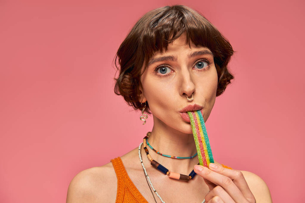 portrait of playful woman in her 20s biting sweet and sour candy strip on pink background - Photo, Image