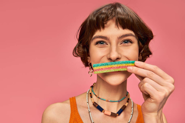 portrait of woman in her 20s covering lips with sweet and sour candy strip on pink background - Photo, Image
