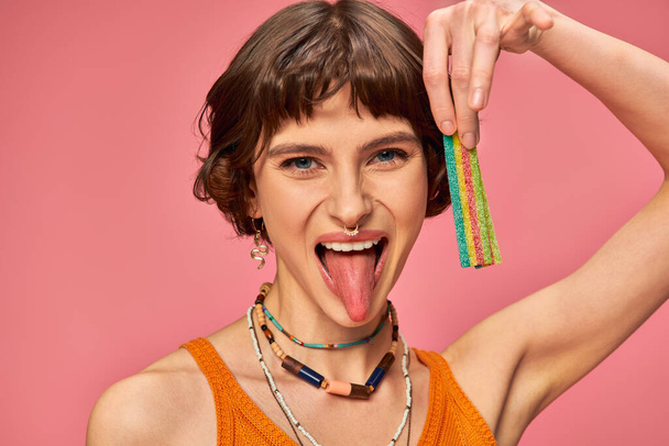 happy woman in her 20s holding sweet and sour candy strip and sticking tongue on pink background - Photo, Image