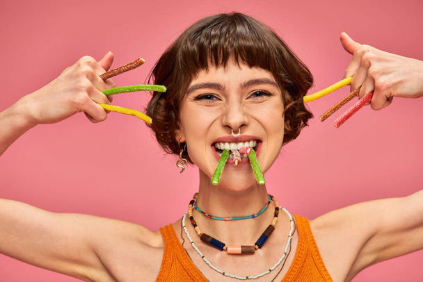 happy young woman in her 20s with sweet and sour candies in hands and teeth on pink background - Photo, Image