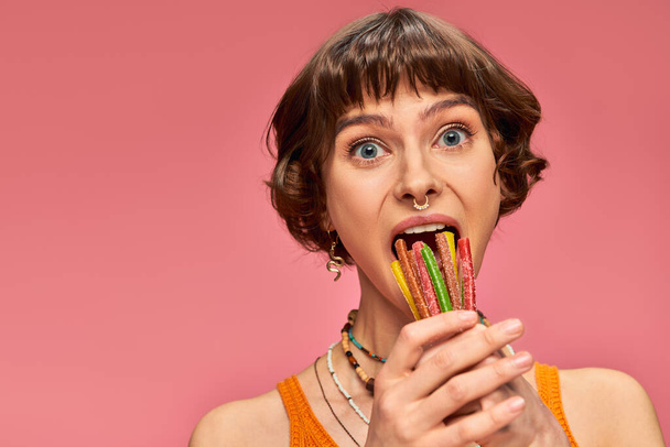 20 'li yaşlarındaki komik genç bir kadın pembe üzerine açık ağzına bir demet tatlı ve ekşi şeker koyuyor. - Fotoğraf, Görsel