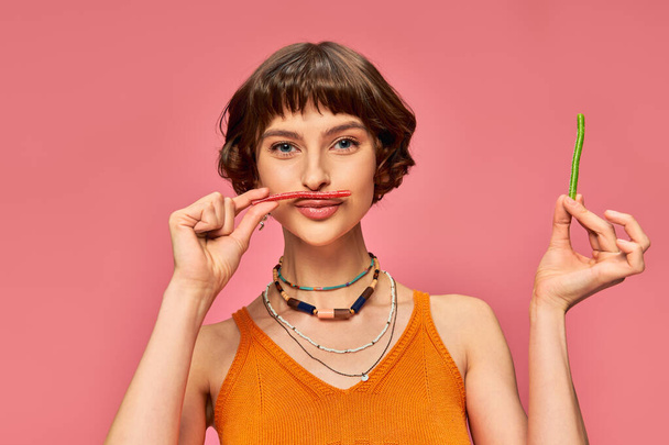playful girl in her 20s smiling and holding sweet and sour candy over her lips and mustache on pink - Fotoğraf, Görsel