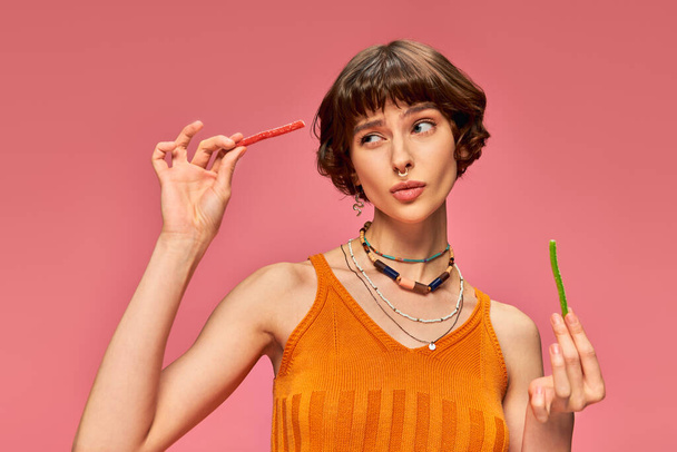 сомнительная девушка с пирсингом в возрасте 20 лет ест два разных вкуса сладких и кислых конфет на розовом - Фото, изображение