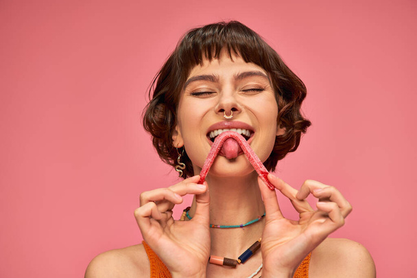 pleased and young woman with nose piercing licking sweet and sour candy strip on pink background - Photo, Image