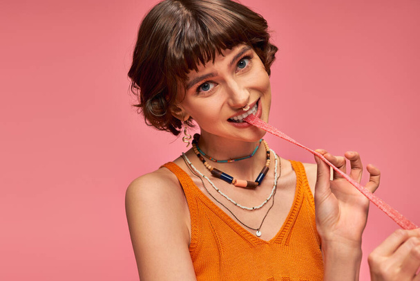 słodkie dziewczyna w 20s z przekłuwanie nosa i białe zęby gryzienie słodko-kwaśny pasek cukierki, różowy zestaw - Zdjęcie, obraz