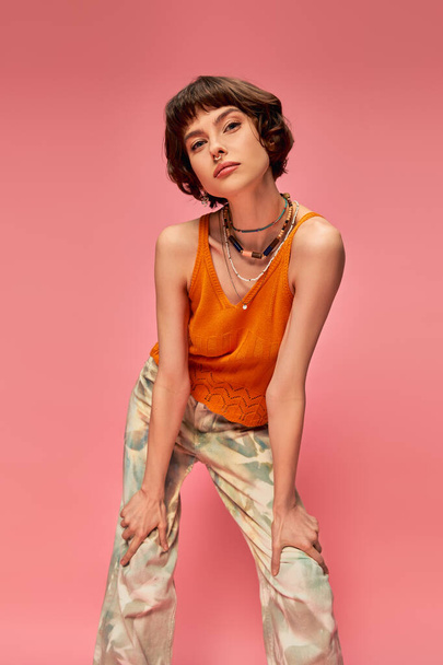 young woman in her 20s with short brunette hair posing in vibrant summer attire on pink backdrop - Photo, Image