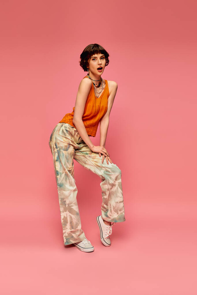 amazed young woman with short brunette hair in vibrant summer attire posing on pink backdrop - Photo, Image
