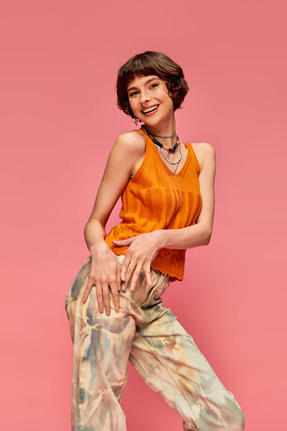 positive young woman with short brunette hair in vibrant summer attire laughing on pink backdrop - Photo, Image