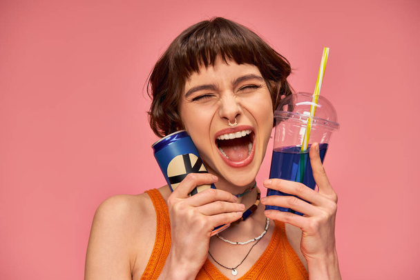 excited young woman with short brunette hair and piercing holding summer drinks on pink backdrop - Photo, Image