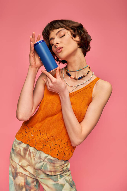 jovem com cabelo curto morena e piercing lábios bebendo perto de lata de refrigerante em rosa, bebida de verão - Foto, Imagem