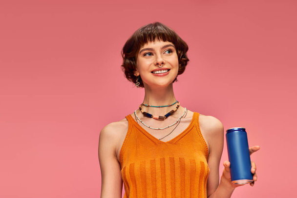 jovem alegre com cabelo morena curta segurando lata de refrigerante enquanto posando em rosa, bebida de verão - Foto, Imagem