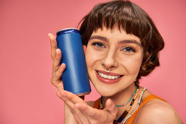 close up of joyful young woman with short brunette hair holding soda can on pink, summer beverage - Photo, Image