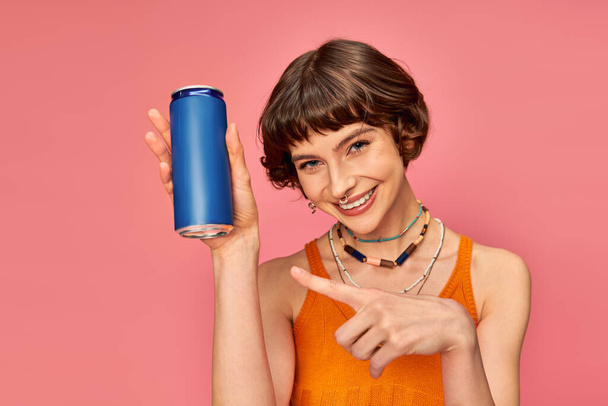 radostná mladá žena s krátkými brunetovými vlasy ukazující na plechovku od limonády na růžové, letní nápoj - Fotografie, Obrázek