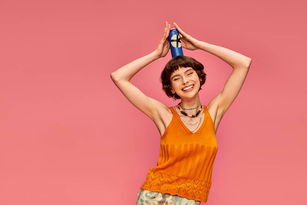 joyful young woman with short brunette hair posing with soda can on her head on pink, summer drink - Photo, Image