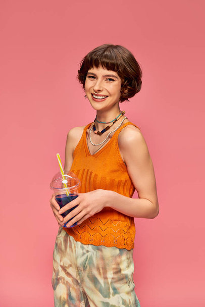 joyeuse jeune femme aux cheveux bruns courts posant avec cocktail d'été rafraîchissant sur rose - Photo, image