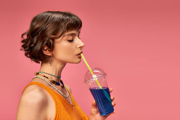 young woman with short brunette hair drinking refreshing summer cocktail from straw on pink - Photo, Image