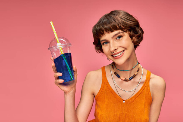 mujer joven positiva con pelo corto morena celebración refrescante bebida de verano en el telón de fondo rosa - Foto, Imagen