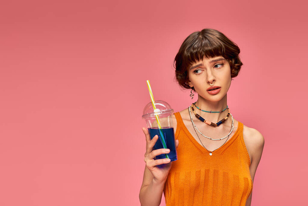 displeased young woman with short brunette hair holding refreshing summer drink on pink backdrop - Photo, Image