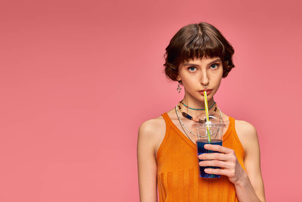 Kısa esmer, genç bir kadın pembe arka planda mavi ferahlatıcı yaz kokteyli içiyor. - Fotoğraf, Görsel