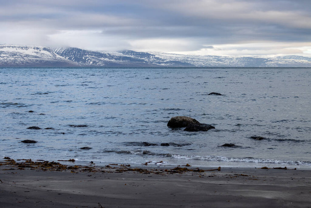 Ciemny wulkaniczny brzeg Atlantyku. Góry na horyzoncie, ze szczytami śniegu. Intensywne zachmurzone niebo. Obszar Isafjordur, Fiordy Zachodnie, Islandia. - Zdjęcie, obraz