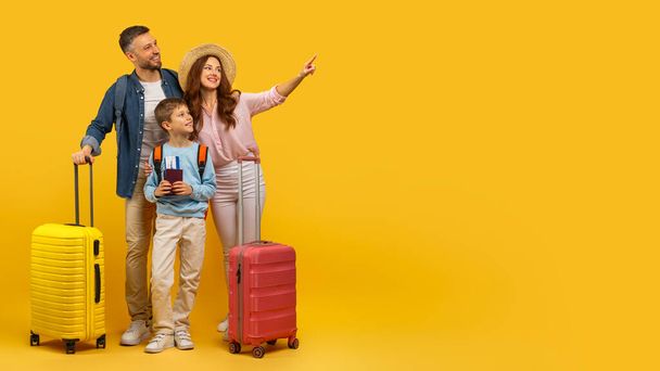 Une famille heureuse avec des valises pointant vers quelque chose, prête pour les vacances, sur un fond jaune vif - Photo, image