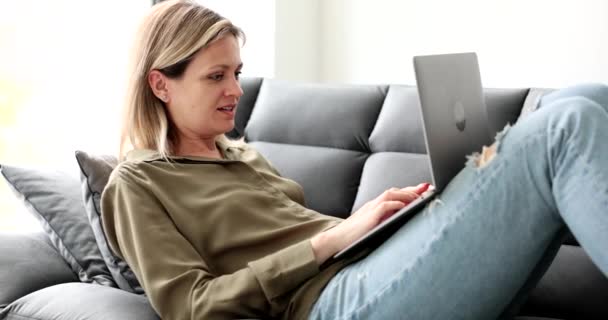 Νεαρή γυναίκα κάθεται στον καναπέ με φορητό υπολογιστή στο γόνατο και κουβέντα σε απευθείας σύνδεση 4k ταινία. Ελεύθερος επαγγελματίας και εργασία εξ αποστάσεως από το σπίτι έννοια - Πλάνα, βίντεο