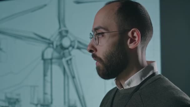 Çift taraflı rüzgar enerjili erkek mühendisin gözlüklü yan çekim görüntüleri ve tabletle birlikte içerideki toplantı sırasında izleyicilere yel değirmeni üretim endüstrisinde gelişmeler sunuluyor. - Video, Çekim