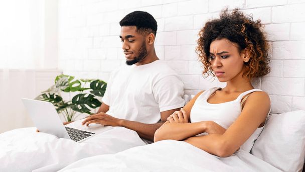 Technologie- und Beziehungsprobleme. Süchtiger junger schwarzer Mann liegt mit Laptop im Bett, Frau sieht wütend und beleidigt aus, leerer Raum - Foto, Bild