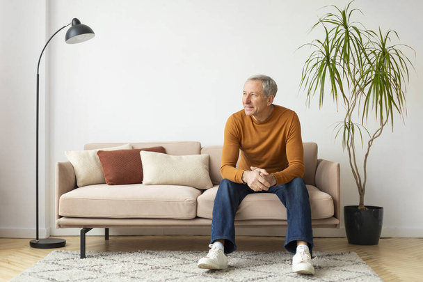 Homme âgé assis confortablement sur un canapé au milieu d'oreillers aux tons chauds, avec une plante en pot en arrière-plan - Photo, image