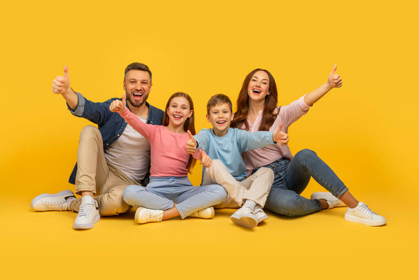 Eine fröhliche vierköpfige Familie mit zwei Kindern und Eltern, die die Daumen nach oben drücken, alle lächelnd auf einem leuchtend gelben Hintergrund - Foto, Bild