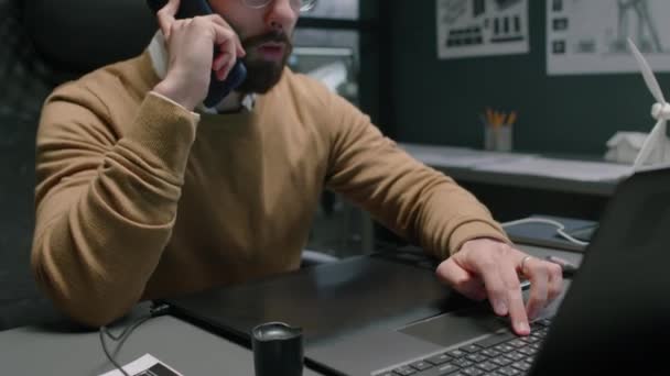 Sakallı, çift taraflı yeşil enerji uzmanının dizüstü bilgisayarla çalışan ve gece ofis masasında otururken akıllı telefonla konuşan görüntüsü. - Video, Çekim