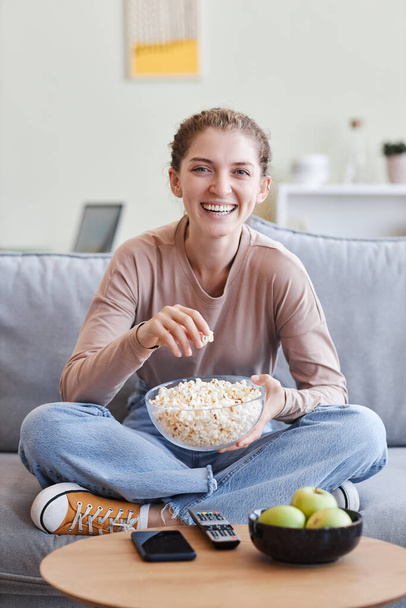 Vertikales Porträt einer unbeschwerten jungen Frau, die mit überkreuzten Beinen auf einem Sofa sitzt und Popcorn in der Hand hält - Foto, Bild