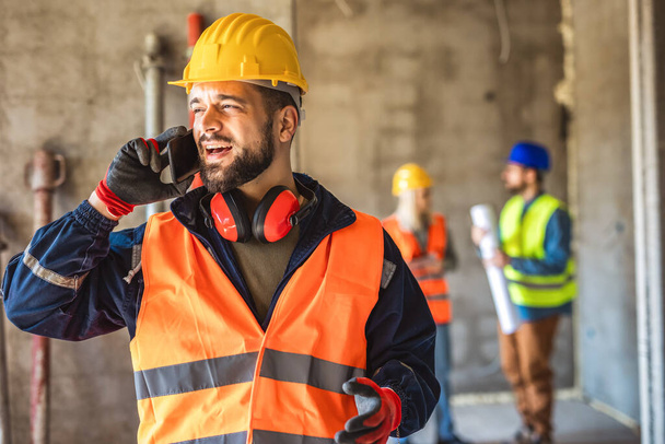 Gestionnaire de chantier debout portant un gilet de sécurité et un casque, parlant au téléphone sur le chantier de construction.  - Photo, image