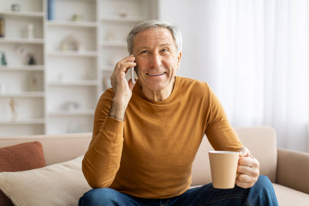 Веселый пожилой мужчина болтает на смартфоне с кофейной кружкой в руке, сидя в хорошо освещенной гостиной - Фото, изображение