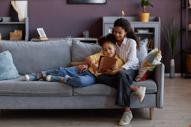 Retrato de cuerpo entero de madre e hija amorosas usando tableta digital juntas mientras se relajan en el sofá en casa, espacio para copiar - Foto, imagen