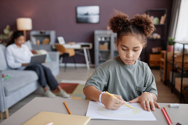 Πρόσοψη πορτρέτο του χαριτωμένο μαύρο κορίτσι ζωγραφίζει εικόνες στο τραπέζι στο σπίτι με τη μητέρα που εργάζονται στο παρασκήνιο, αντίγραφο χώρο - Φωτογραφία, εικόνα