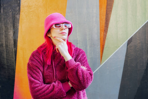 Hipster young woman with pink hair and sunglasses in magenta fluffy sweatshirt and bucket hat posing on the wall background. Urban street fashion. Mono color look. Gen Z, millennials self expression - Фото, зображення