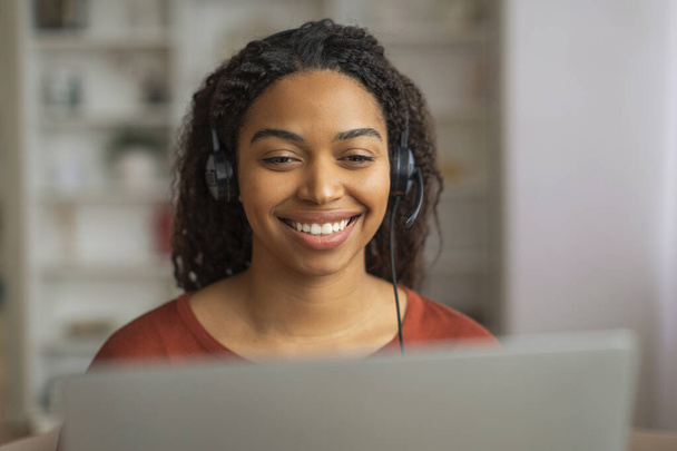 Uma mulher negra alegre em um top vermelho e fone de ouvido envolvente com seu laptop, mulher afro-americana desfrutando de uma conversa on-line ou entretenimento multimídia, trabalhando como operadora de call center - Foto, Imagem