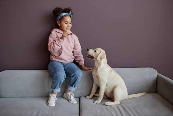 Full length portrait of black little girl playing with dog indoors against purple wall, copy space - Photo, Image