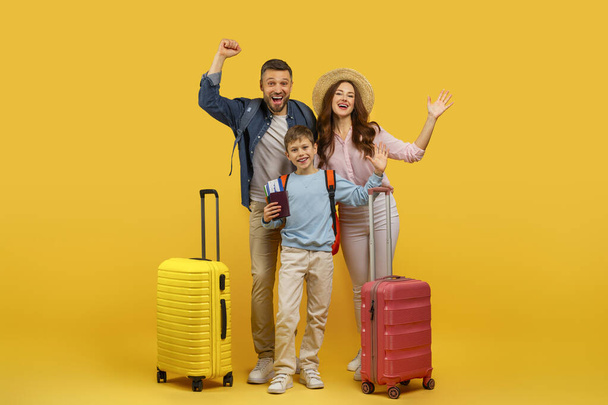 Μια χαρούμενη οικογένεια με βαλίτσες που ποζάρουν με ενθουσιασμό σε κίτρινο φόντο, επιδεικνύοντας ενθουσιασμό για ταξίδια - Φωτογραφία, εικόνα
