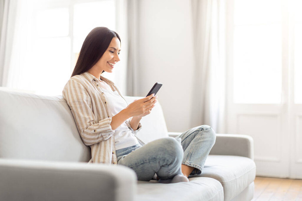 Hymyilevä nainen, joka istuu sohvalla, pitää älypuhelinta, huokuu mukavuutta ja helppoutta kotona, kopioi tilaa - Valokuva, kuva
