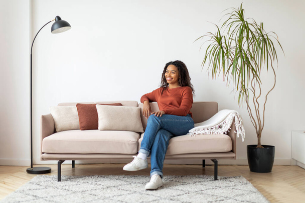 Onnellinen musta nainen istuu rennosti sohvalla, ympäröi minimalistinen sisustus, nauttia rauhallinen hetki. - Valokuva, kuva