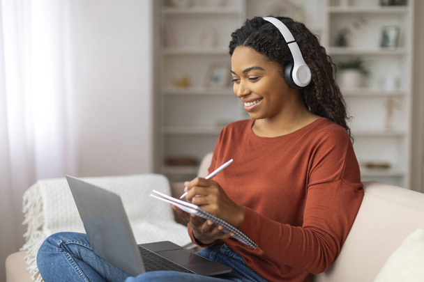 Усміхнена жінка сидить на дивані, використовуючи ноутбук, одягаючи навушники, щаслива афроамериканська жінка приймає нотатки в блокнот, відвідуючи онлайн-урок - Фото, зображення
