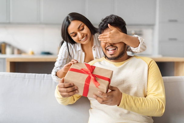 A joyous indian woman surprising a delighted man by covering his eyes and offering a gift with a red ribbon. - Photo, Image
