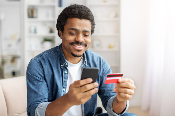 Wesoły czarny mężczyzna w luźnym stroju zajmuje się swoim smartfonem trzymając kartę kredytową, potencjalnie zarządzając finansami lub robiąc zakupy online - Zdjęcie, obraz