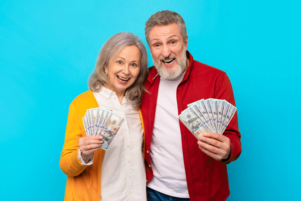 Eläkevauraus. Hymyilevä vanhempi mies ja nainen pitämässä dollarin rahaa käteisenä sinistä studiotaustaa vasten, jakamassa voiton ja menestyksen jännitystä. Taloudellinen turvallisuus ja varallisuus - Valokuva, kuva