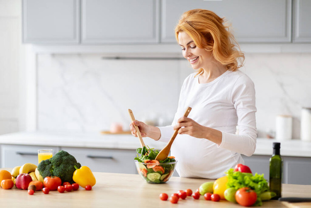 Una donna bionda incinta ha visto mescolare un'insalata, circondata da verdure fresche e crude in una cucina moderna - Foto, immagini