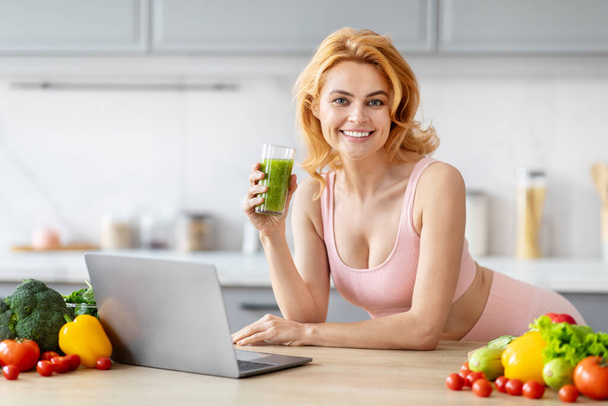 Fit blonde woman enjoying a green smoothie while working on a laptop in an organized kitchen - Photo, Image